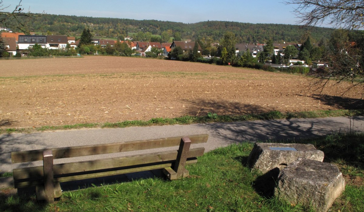 Eine Holzbank steht vor einem Feld, dahinter sind H&auml;user und ein bewaldeter H&uuml;gel zu sehen. Der Himmel ist klar und blau., &copy; RadL Leonberg
