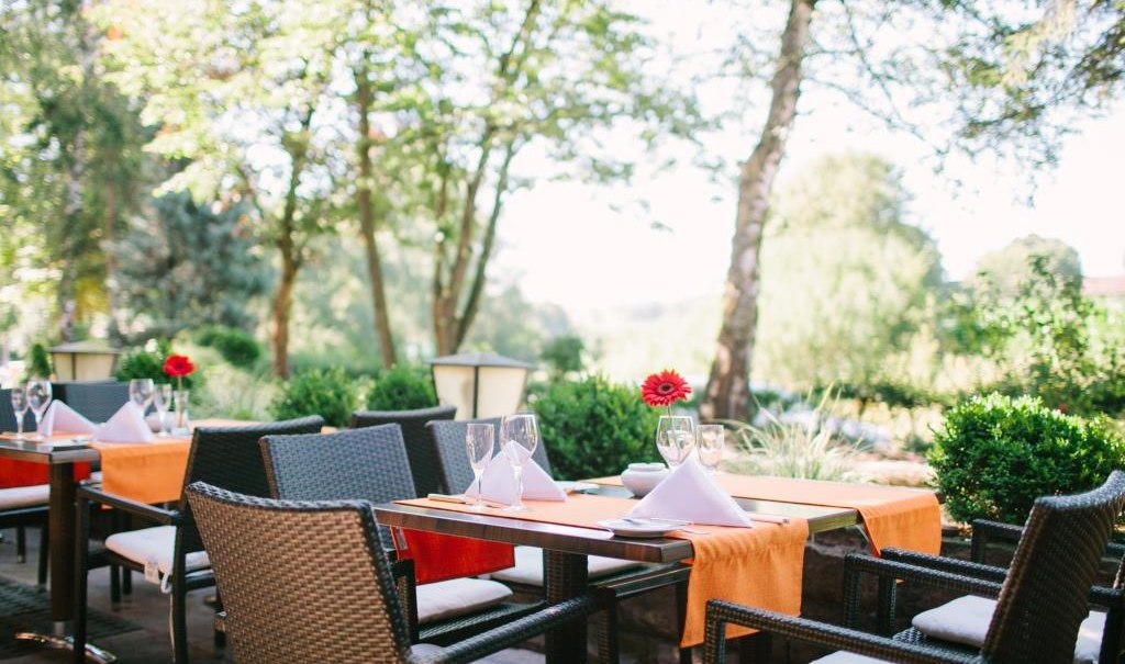 Gedeckte Tische auf einer Terrasse mit orangefarbenen Tischläufern und roten Blumen, umgeben von Bäumen und Sträuchern., © relexa Waldhotel Schatten Gedeckte Tische auf einer Terrasse mit orangefarbenen Tischläufern und roten Blumen, umgeben von Bäumen und Sträuchern., © relexa Waldhotel Schatten