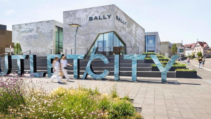 Große Buchstaben formen das Wort 'OUTLETCITY' vor modernen Gebäuden. Menschen gehen vorbei, während Blumen im Vordergrund blühen., © OUTLETCITY AG Große Buchstaben formen das Wort 'OUTLETCITY' vor modernen Gebäuden. Menschen gehen vorbei, während Blumen im Vordergrund blühen., © OUTLETCITY AG