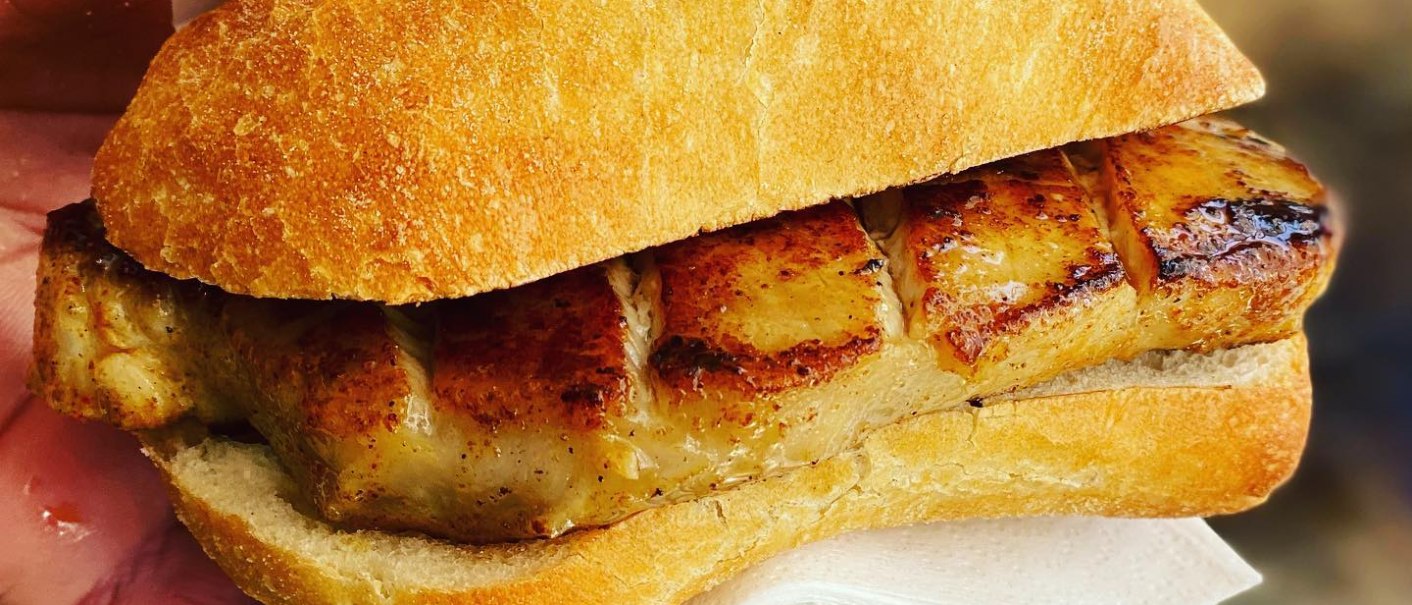 Ein knuspriges Sandwich mit gegrilltem Fleisch, das in einer Hand gehalten wird. Eine Serviette ist zwischen Hand und Sandwich gelegt., © Salva's Curry-Truck