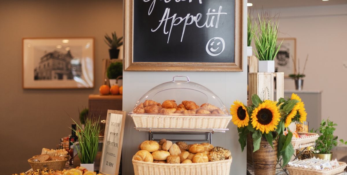 Ein Frühstücksbuffet mit Brötchen, Croissants und Sonnenblumen. Ein Schild mit 'Guten Appetit' hängt darüber. Pflanzen und Gebäck sind dekorativ arrangiert., © Novum Hospitality