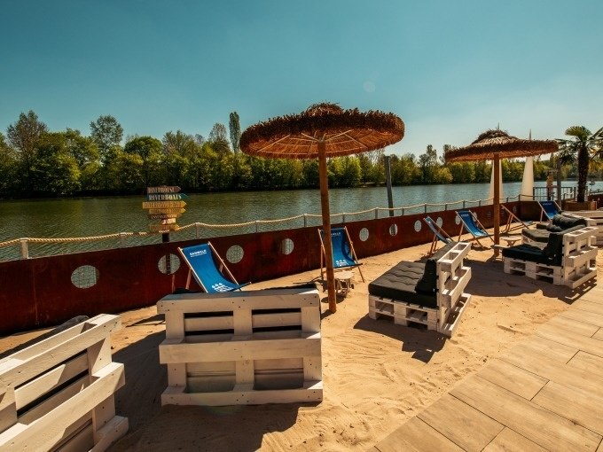 Strandbereich mit Palettenmöbeln, Sonnenschirmen und Liegestühlen am Flussufer, umgeben von Bäumen und klarem Himmel., © Cool-Tours StattReisen Strandbereich mit Palettenmöbeln, Sonnenschirmen und Liegestühlen am Flussufer, umgeben von Bäumen und klarem Himmel., © Cool-Tours StattReisen