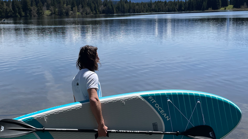 Eine Person steht am Ufer eines Sees mit einem SUP-Board und Paddel, umgeben von Wald und ruhigem Wasser., © seezeit GmbH