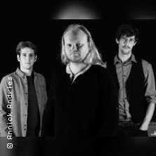 Drei M&auml;nner posieren in Schwarz-Wei&szlig;. Der Mann in der Mitte hat lange Haare und tr&auml;gt ein dunkles Oberteil. Die anderen stehen leicht im Hintergrund., &copy; links im Bild