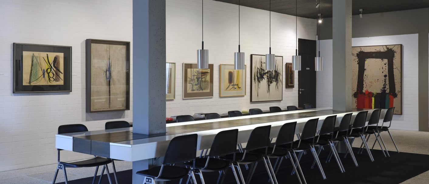 Moderner Konferenzraum mit langen Tisch, schwarzen Stühlen und abstrakten Kunstwerken an weißen Wänden. Hängelampen beleuchten den Raum., © Volker Naumann Moderner Konferenzraum mit langen Tisch, schwarzen Stühlen und abstrakten Kunstwerken an weißen Wänden. Hängelampen beleuchten den Raum., © Volker Naumann