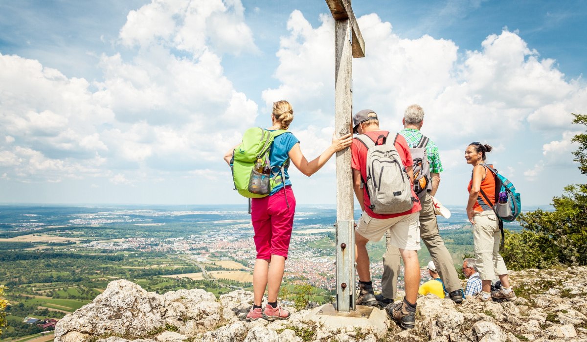 Wanderer stehen auf einem Berggipfel neben einem Gipfelkreuz und genießen die Aussicht auf die Landschaft unter einem blauen Himmel mit Wolken., © hochgehberge