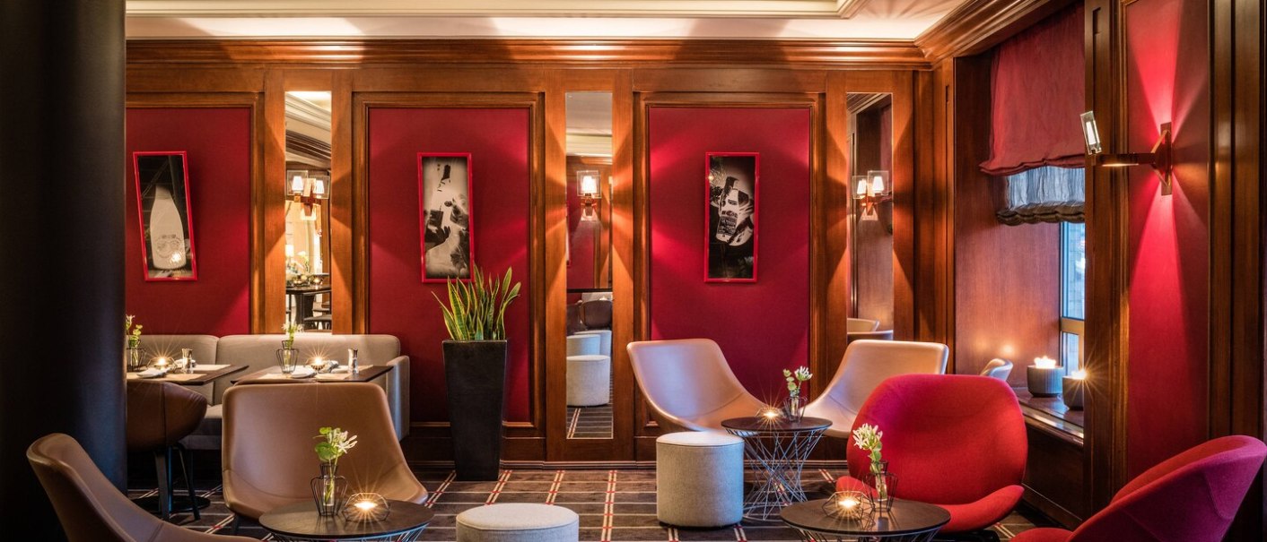 Elegante Lounge im Le Méridien Stuttgart mit roten Wänden, modernen Sesseln und kleinen Tischen. Dezente Beleuchtung und stilvolle Dekoration., © TOMAS