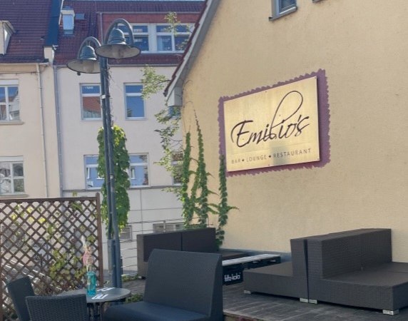 Au&szlig;enbereich von Emilio's Bar, Lounge und Restaurant mit modernen Sitzm&ouml;beln und Pflanzen. Ein Schild mit dem Namen des Lokals ist an der Wand angebracht., &copy; Emilio's