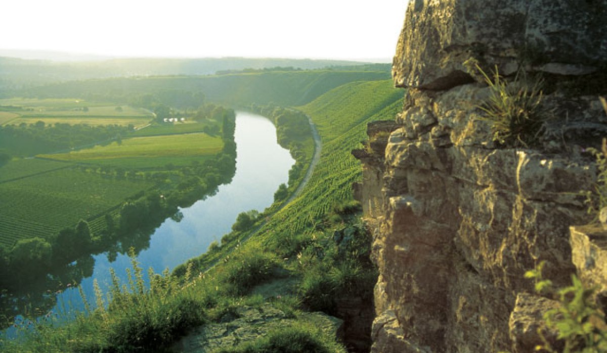 Ein Fluss schlängelt sich durch eine grüne Landschaft, umgeben von Feldern und Hügeln. Im Vordergrund sind Felsen zu sehen., © Land der 1000 Hügel - Kraichgau-Stromberg Ein Fluss schlängelt sich durch eine grüne Landschaft, umgeben von Feldern und Hügeln. Im Vordergrund sind Felsen zu sehen., © Land der 1000 Hügel - Kraichgau-Stromberg