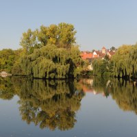 Ein ruhiger Fluss mit üppigen Bäumen und roten Dächern im Hintergrund, die sich im klaren Wasser spiegeln., © Land der 1000 Hügel - Kraichgau-Stromberg Ein ruhiger Fluss mit üppigen Bäumen und roten Dächern im Hintergrund, die sich im klaren Wasser spiegeln., © Land der 1000 Hügel - Kraichgau-Stromberg