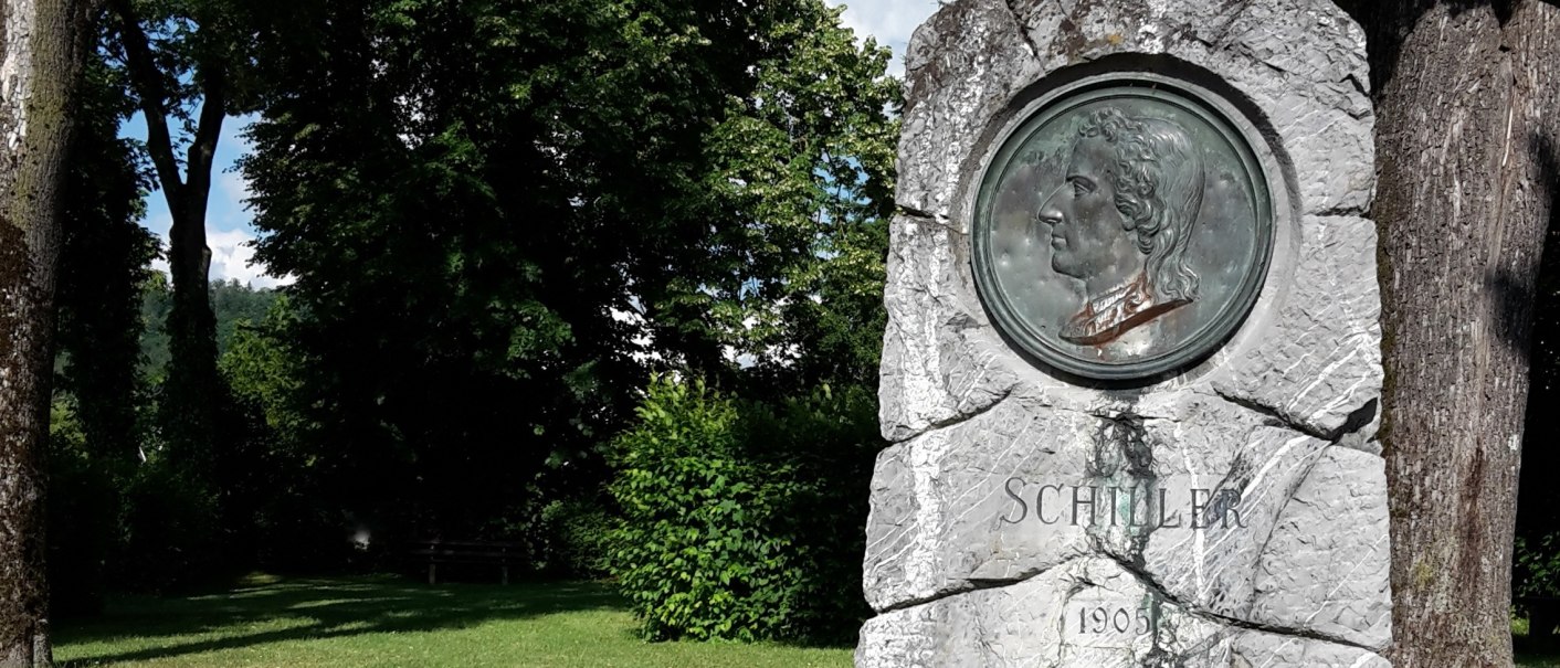 Schillerdenkmal in grüner Parkanlage mit Büschen und Bäumen - grob behauene, graue Natursteinsäule, mit bronzenem Portät-Medaillon, darunter die Inschrift "Schiller" und die Jahreszahl 1905, © Petra Natzkowski Schillerdenkmal in grüner Parkanlage mit Büschen und Bäumen - grob behauene, graue Natursteinsäule, mit bronzenem Portät-Medaillon, darunter die Inschrift "Schiller" und die Jahreszahl 1905, © Petra Natzkowski