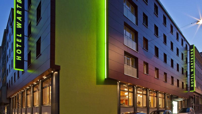 Moderne Hotelaußenansicht bei Dämmerung, mit grüner Beleuchtung und parkenden Autos. Der Schriftzug 'Hotel Wartburg' ist sichtbar., © VCH Hotel Wartburg Moderne Hotelaußenansicht bei Dämmerung, mit grüner Beleuchtung und parkenden Autos. Der Schriftzug 'Hotel Wartburg' ist sichtbar., © VCH Hotel Wartburg