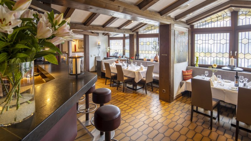 Ein gemütliches Restaurant mit Holzbalkendecke, gedeckten Tischen und großen Fenstern. Blumen und Kerzen sorgen für eine einladende Atmosphäre., © Hotel Gloria GmbH