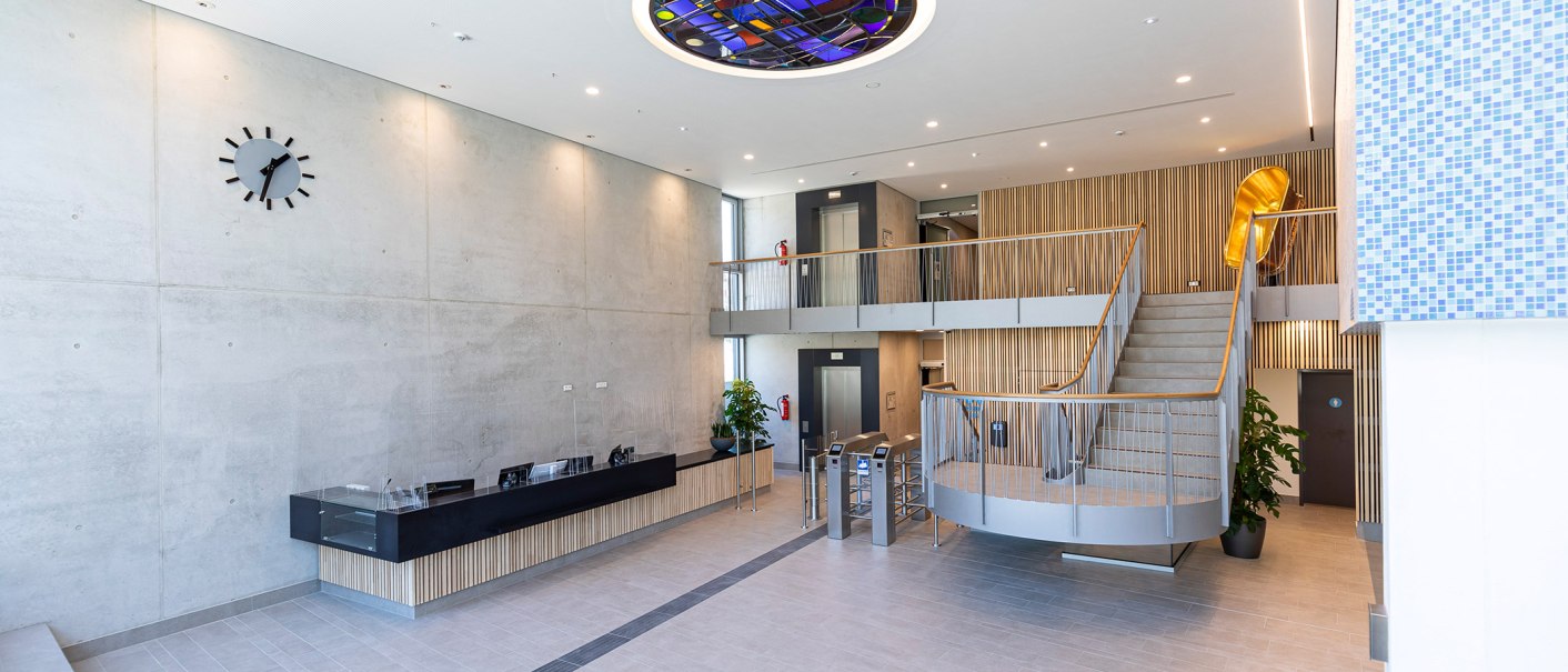 Modernes Foyer mit Empfangstresen, Treppe, Aufzügen und einer großen Wanduhr. Die Decke hat ein buntes Glaselement., © Stuttgarter Bäder