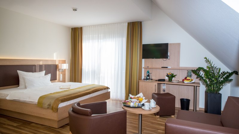 Modernes Doppelzimmer mit großem Bett, Tisch, zwei Sesseln, TV und Pflanzen. Helle Vorhänge und Holzmöbel schaffen eine gemütliche Atmosphäre., © benjamin stollenberg | fotografie