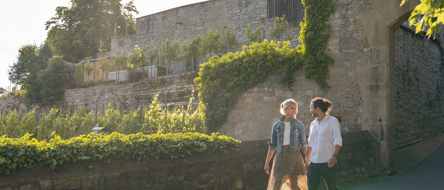 Ein Paar spaziert an einer alten Steinmauer entlang, umgeben von grünen Pflanzen und Weinreben, bei sonnigem Wetter., © Stuttgart-Marketing GmbH, Martina Denker Ein Paar spaziert an einer alten Steinmauer entlang, umgeben von grünen Pflanzen und Weinreben, bei sonnigem Wetter., © Stuttgart-Marketing GmbH, Martina Denker