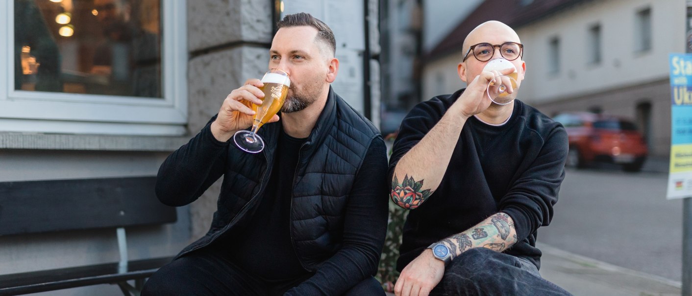 Zwei Männer sitzen auf einer Holzpalette vor einem Gebäude und trinken Bier. Einer trägt eine Brille und hat tätowierte Arme., © Kraftpaule GmbH