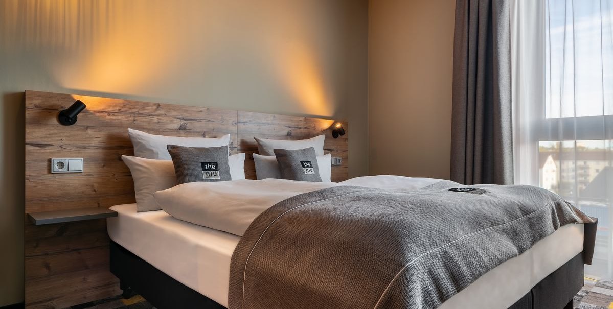 Modernes Hotelzimmer mit Doppelbett, Holzkopfteil, grauen Kissen und Vorhängen. Warmes Licht beleuchtet den Raum., © Novum Hospitality Modernes Hotelzimmer mit Doppelbett, Holzkopfteil, grauen Kissen und Vorhängen. Warmes Licht beleuchtet den Raum., © Novum Hospitality