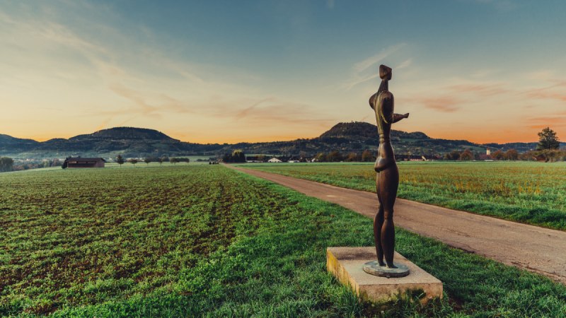 Eine Skulptur steht auf einem Feldweg, umgeben von grünen Feldern und Hügeln im Hintergrund, während die Sonne untergeht., © Landratsamt Ostalbkreis