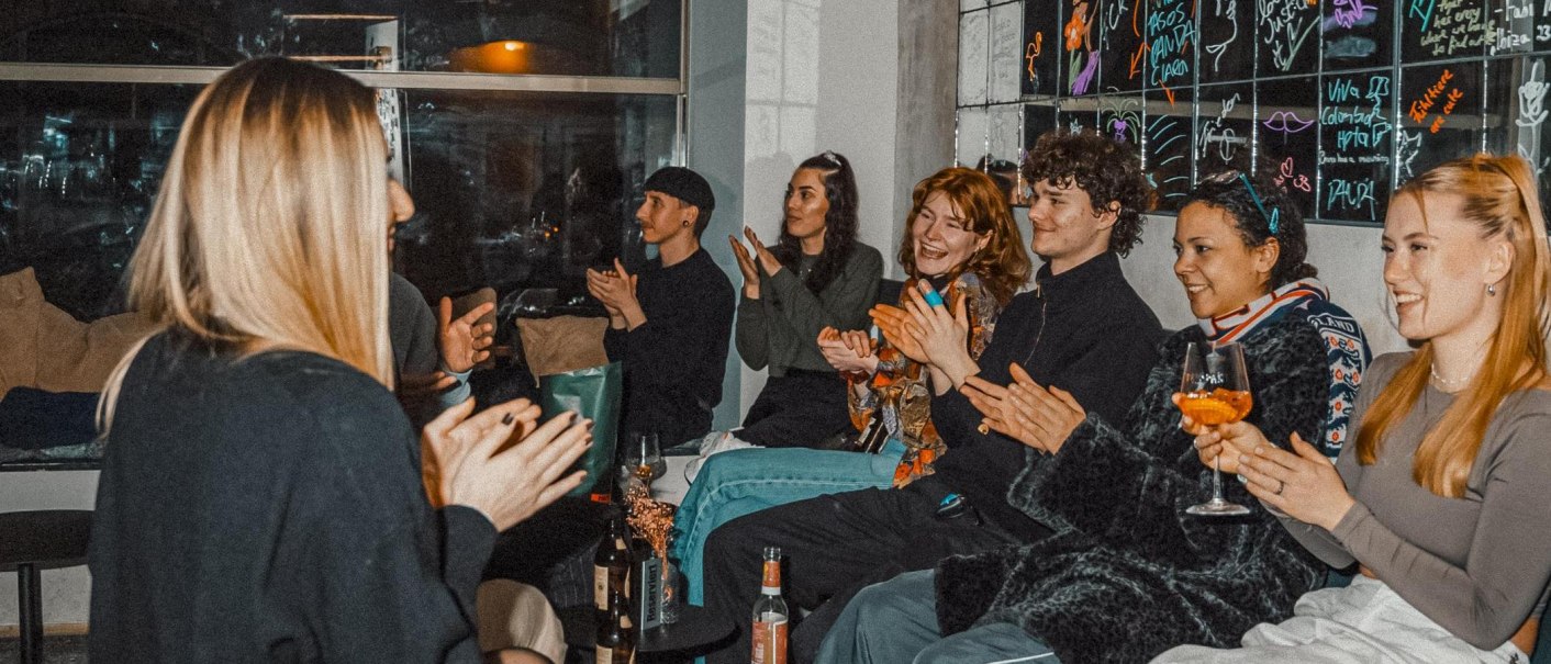 Eine Gruppe von Menschen sitzt in einem Raum, klatscht und lacht. Einige halten Getränke in der Hand. Im Hintergrund sind bunte Kritzeleien an der Wand., © Bruno, Stuttgart Eine Gruppe von Menschen sitzt in einem Raum, klatscht und lacht. Einige halten Getränke in der Hand. Im Hintergrund sind bunte Kritzeleien an der Wand., © Bruno, Stuttgart
