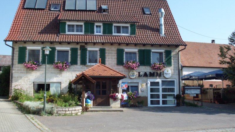 Ein traditionelles Gasthaus mit roten Dachziegeln, gr&uuml;nen Fensterl&auml;den und Blumenk&auml;sten. Ein Biergarten ist rechts zu sehen.