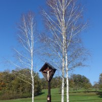 Ein Kruzifix steht zwischen zwei kahlen Birken auf einer grünen Wiese unter klarem, blauem Himmel., © Foto: Cornelia Steinbach