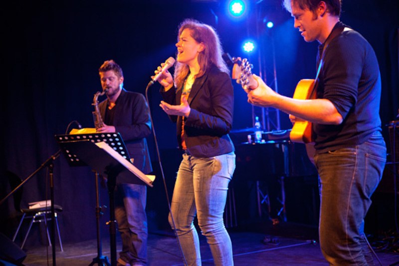 Drei Musiker auf der Bühne: Eine Frau singt, begleitet von einem Gitarristen und einem Saxophonisten. Die Bühne ist in blaues Licht getaucht., © Jazzclub Schwäbisch Hall e.V.