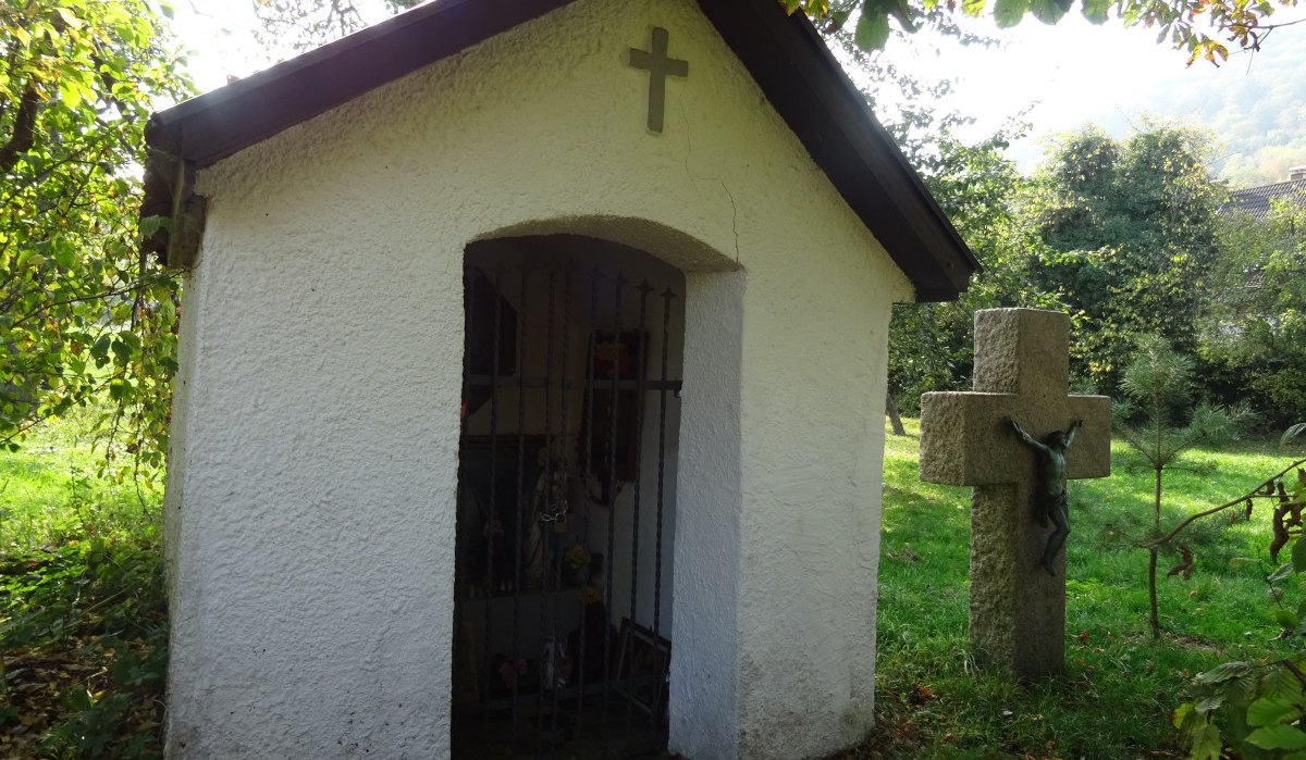 Weiße Kapelle mit Kreuz und Gittertür, daneben Steinkreuz mit Christusfigur, umgeben von Bäumen und Gras., © Foto: Cornelia Steinbach Weiße Kapelle mit Kreuz und Gittertür, daneben Steinkreuz mit Christusfigur, umgeben von Bäumen und Gras., © Foto: Cornelia Steinbach