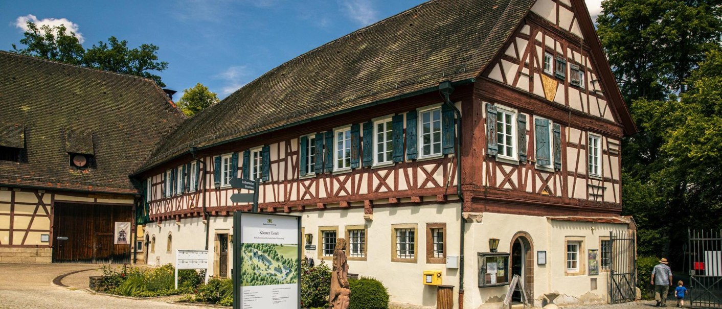 Fachwerkgebäude des Klosters Lorch mit blauen Fensterläden. Ein Informationsschild steht im Vordergrund, umgeben von Bäumen und einem klaren Himmel., © Stuttgart-Marketing GmbH, Sarah Schmid Fachwerkgebäude des Klosters Lorch mit blauen Fensterläden. Ein Informationsschild steht im Vordergrund, umgeben von Bäumen und einem klaren Himmel., © Stuttgart-Marketing GmbH, Sarah Schmid