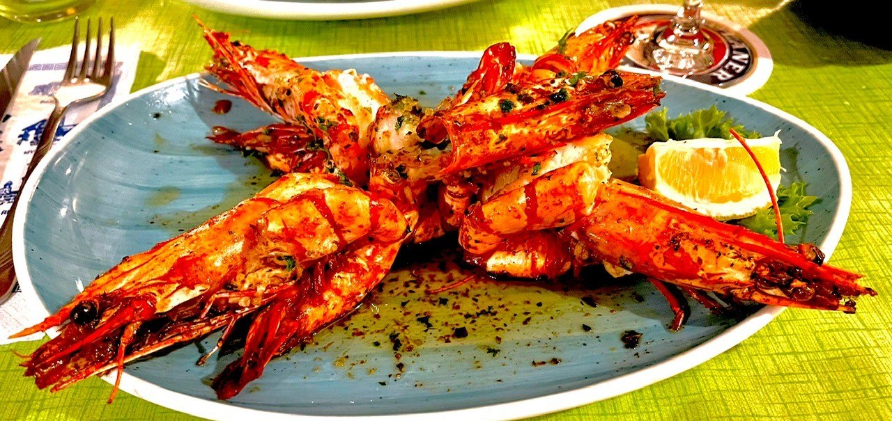 Gegrillte Garnelen auf blauem Teller mit Kräutern und Zitronenscheibe, auf grünem Tisch serviert., © Restaurant Neckarau, P. Kotaidis