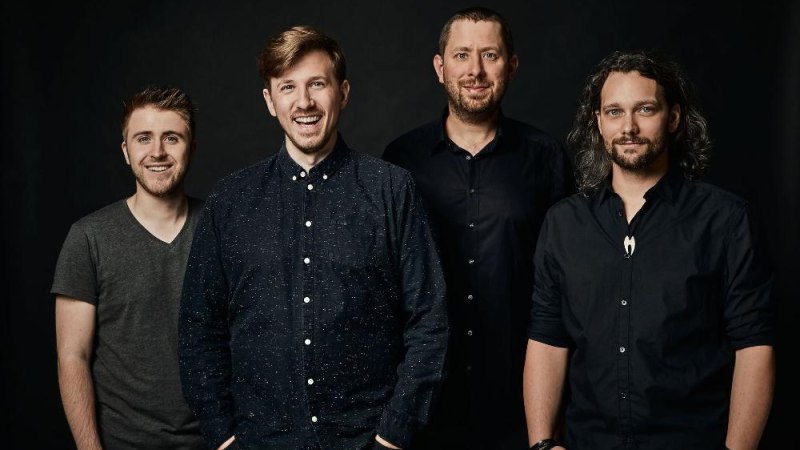 Vier Männer in dunkler Kleidung lächeln vor einem schwarzen Hintergrund in die Kamera., © Foto Alexander Wunsch Vier Männer in dunkler Kleidung lächeln vor einem schwarzen Hintergrund in die Kamera., © Foto Alexander Wunsch