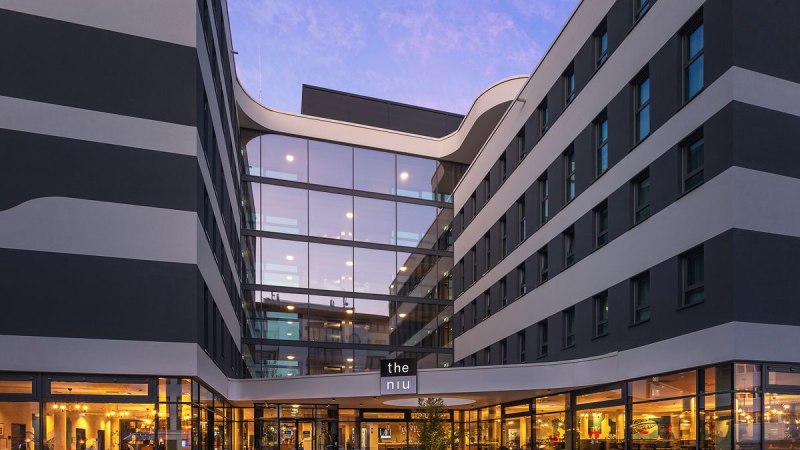 Moderne Hotelaußenfassade mit Glasfront und beleuchtetem Eingangsbereich bei Dämmerung. Das Gebäude hat ein elegantes, wellenförmiges Design., © Novum Hospitality Moderne Hotelaußenfassade mit Glasfront und beleuchtetem Eingangsbereich bei Dämmerung. Das Gebäude hat ein elegantes, wellenförmiges Design., © Novum Hospitality