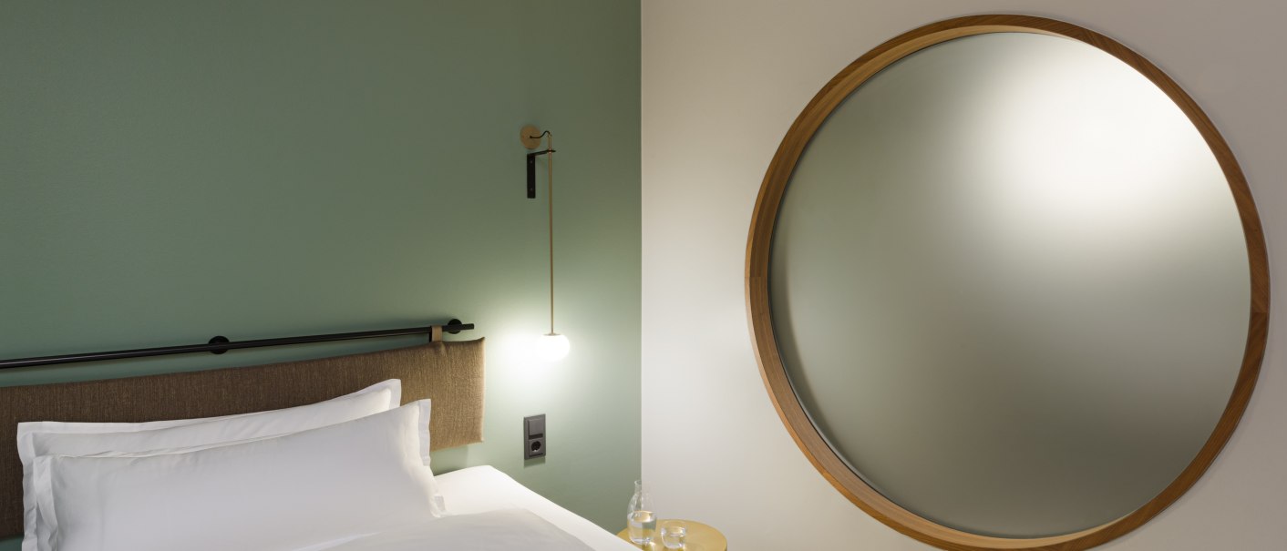 Ein Bett mit wei&szlig;er Bettw&auml;sche, eine runde Wandlampe und ein gro&szlig;er runder Spiegel an einer gr&uuml;nen Wand., &copy; EmiLu Hotel GmbH