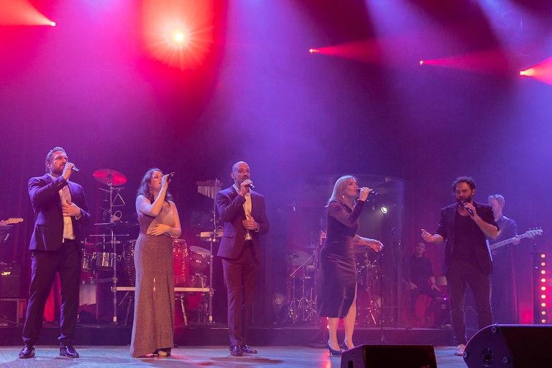 Fünf Sänger performen auf einer Bühne, umgeben von bunten Lichtern. Eine Band spielt im Hintergrund. Die Atmosphäre ist festlich und lebendig., © Schönblick gemeinnützige GmbH Fünf Sänger performen auf einer Bühne, umgeben von bunten Lichtern. Eine Band spielt im Hintergrund. Die Atmosphäre ist festlich und lebendig., © Schönblick gemeinnützige GmbH