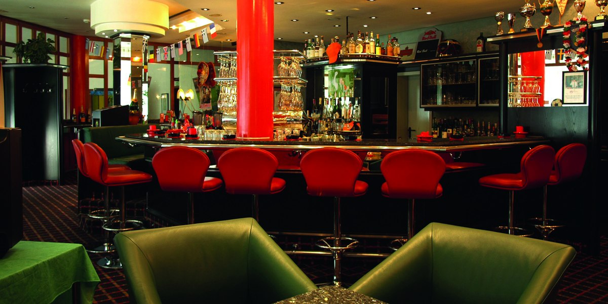 Elegante Bar mit roten Hockern, zentraler Theke und dekorativen Flaschen. Internationale Flaggen und Trophäen schmücken den Raum., © © H-Hotels.com