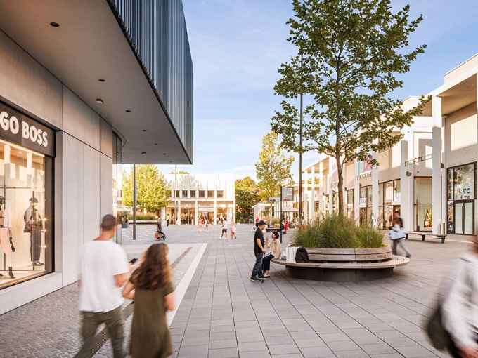 Menschen flanieren durch die moderne Fu&szlig;g&auml;ngerzone der Outletcity Metzingen, umgeben von Gesch&auml;ften und B&auml;umen., &copy; Schlienz-Tours GmbH & Co.KG