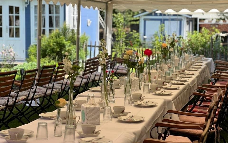 Ein langer Tisch unter einem Pavillon, festlich gedeckt mit Blumenvasen und Geschirr, vor einem blauen Geb&auml;ude im Garten., &copy; cCaf&eacute;Pl&auml;tschwiesle