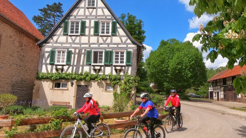 Drei Radfahrer in bunter Kleidung fahren an einem malerischen Fachwerkhaus vorbei. Der Himmel ist blau mit weißen Wolken., © Land der 1000 Hügel - Kraichgau-Stromberg