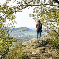 Eine Person steht auf dem Beurener Fels, umgeben von Bäumen, und blickt auf die weite Landschaft von Beuren., © SMG, Sarah Schmid Eine Person steht auf dem Beurener Fels, umgeben von Bäumen, und blickt auf die weite Landschaft von Beuren., © SMG, Sarah Schmid