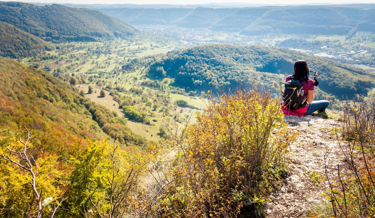 Eine Person sitzt auf einem Hügel und blickt ins Lenninger Tal. Die Landschaft ist von bewaldeten Hügeln und weiten Wiesen geprägt., © hochgehberge