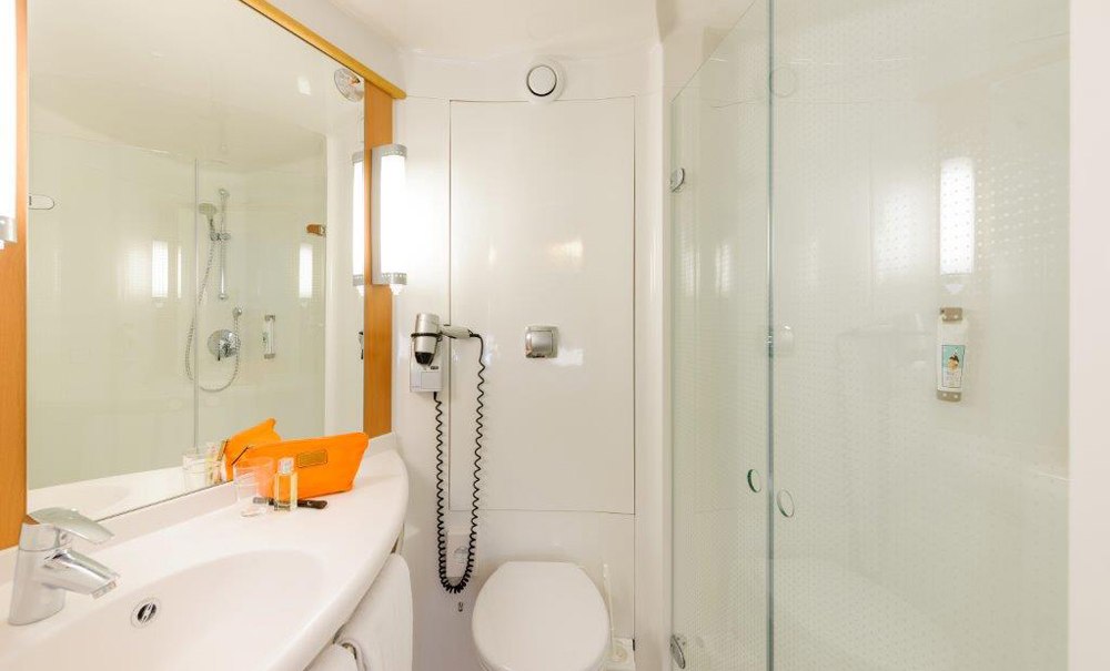 Modernes Badezimmer mit Glasdusche, Waschbecken, großem Spiegel und Toilette. Ein Föhn hängt an der Wand, und eine orangefarbene Tasche steht auf dem Waschbecken., © Ibis Stuttgart Airport Messe Modernes Badezimmer mit Glasdusche, Waschbecken, großem Spiegel und Toilette. Ein Föhn hängt an der Wand, und eine orangefarbene Tasche steht auf dem Waschbecken., © Ibis Stuttgart Airport Messe