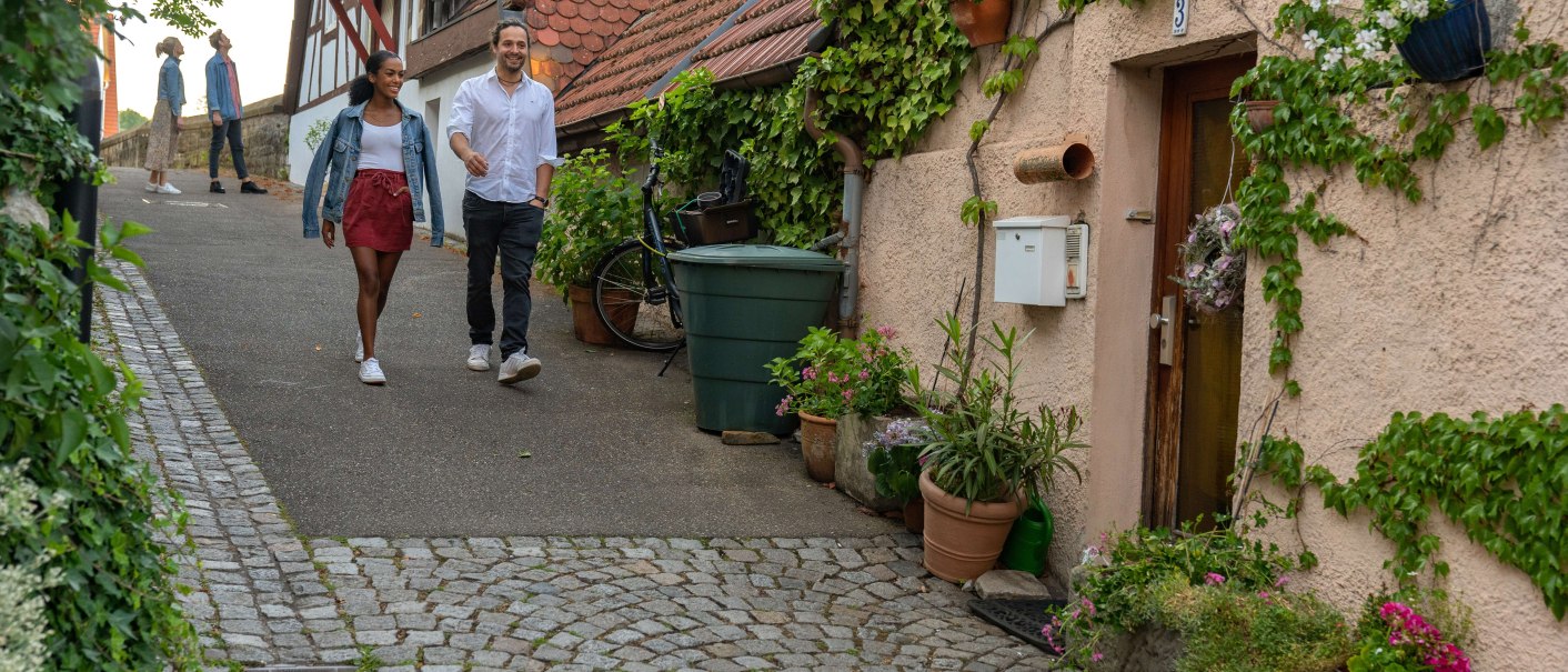Ein Paar spaziert durch eine malerische Altstadtstraße in Besigheim. Fachwerkhäuser und Pflanzen schmücken den Weg. Weitere Personen im Hintergrund., © SMG, Martina Denker Ein Paar spaziert durch eine malerische Altstadtstraße in Besigheim. Fachwerkhäuser und Pflanzen schmücken den Weg. Weitere Personen im Hintergrund., © SMG, Martina Denker
