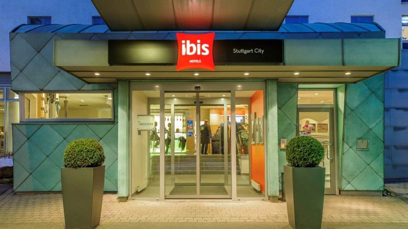 Eingang des Ibis Hotels Stuttgart City mit beleuchtetem Schild und Glasdreht&uuml;r, flankiert von zwei Pflanzen in modernen Pflanzgef&auml;&szlig;en., &copy; TOMAS