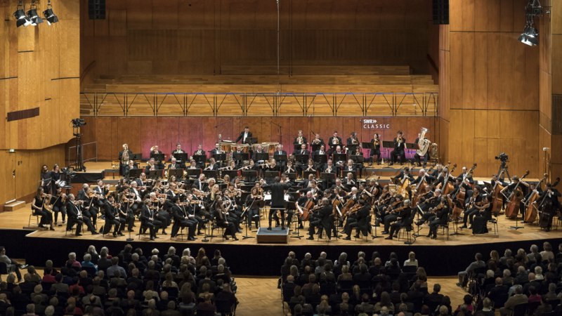 Das SWR Symphonieorchester spielt auf einer großen Bühne vor Publikum. Die Musiker tragen formelle Kleidung, und der Dirigent steht in der Mitte., © SWR Alexander Kluge