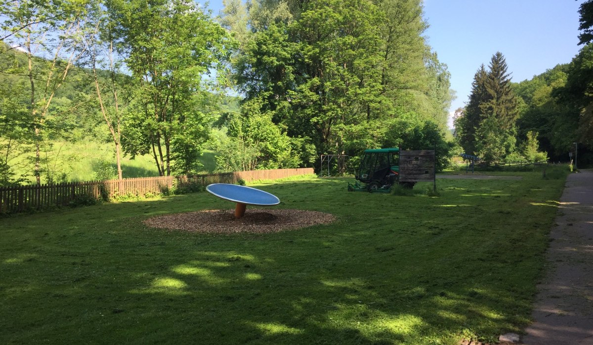Ein grüner Spielplatz mit einer Wippe in der Mitte, umgeben von Bäumen und einem Holzzaun. Ein kleiner Traktor steht im Hintergrund., © www.pro-cycl.de Ein grüner Spielplatz mit einer Wippe in der Mitte, umgeben von Bäumen und einem Holzzaun. Ein kleiner Traktor steht im Hintergrund., © www.pro-cycl.de