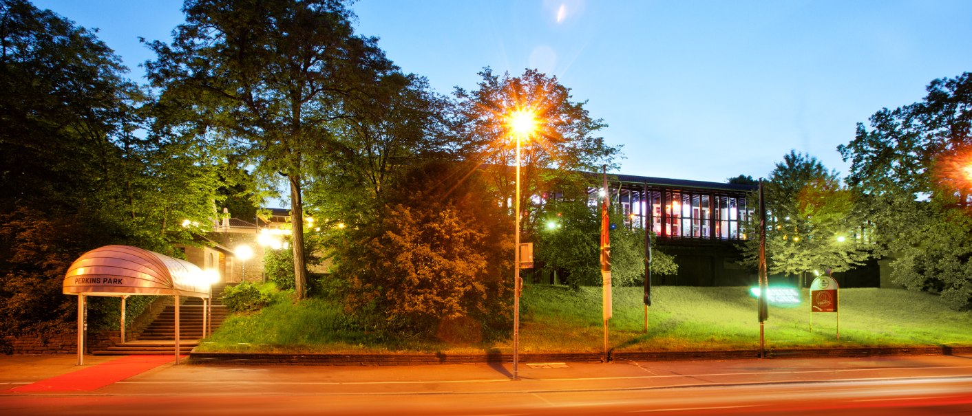 Eingang des Perkins Park bei Nacht, beleuchtet mit rotem Teppich. Im Hintergrund sind Bäume und ein modernes Gebäude zu sehen., © PerkinsPark Eingang des Perkins Park bei Nacht, beleuchtet mit rotem Teppich. Im Hintergrund sind Bäume und ein modernes Gebäude zu sehen., © PerkinsPark