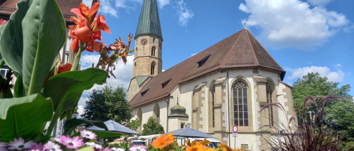 Kirche mit Kirchturm vor blauem Himmel mit weißen Wolken, davor bunte Sommerblumen, © Petra Natzkowski