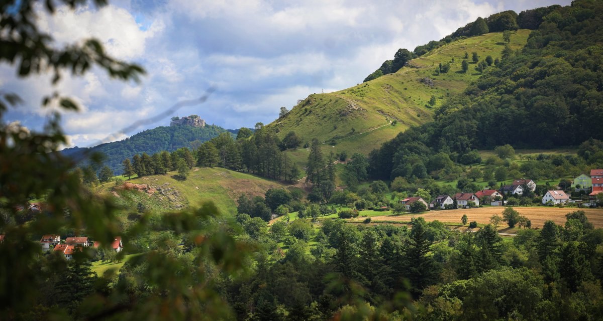 Grüne Hügel mit Bäumen und Häusern, Burgruine auf einem Hügel im Hintergrund, bewölkter Himmel. Grüne Hügel mit Bäumen und Häusern, Burgruine auf einem Hügel im Hintergrund, bewölkter Himmel.