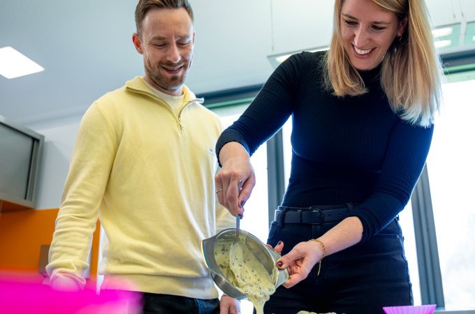 Zwei Personen gießen gemeinsam Schokolade in eine Form. Beide lächeln und wirken konzentriert. Im Hintergrund sind Fenster zu sehen., © Alfred Ritter GmbH &amp; Co. KG | Waldenbuch