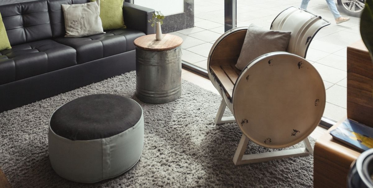 Modernes Wohnzimmer mit schwarzem Sofa, rundem Hocker, Metalltisch und Fassstuhl. Große Fensterfront mit Blick auf die Straße., © Novum Hospitality